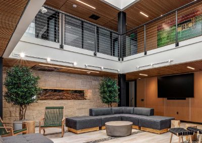 Modern office lobby with gray sectional sofas, a round table, indoor plants, and a TV on a wooden wall.