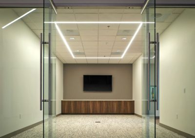 Empty conference room with glass doors, a wall-mounted TV, wood cabinet, and ceiling lights.