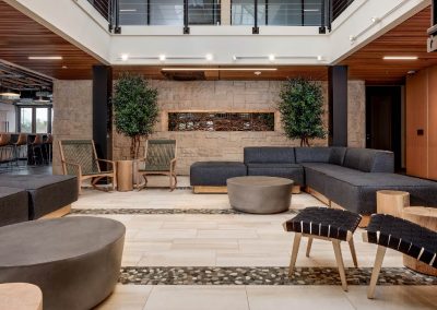 Modern lobby with gray sofas, round tables, plants, and wooden ceiling accents.