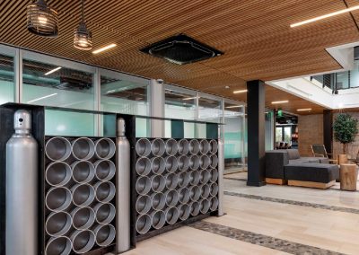 Modern lobby with industrial decor, featuring cylinder display, wooden ceiling, gray seating, and green glass walls.