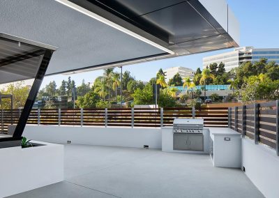 Modern rooftop patio with a barbecue grill, surrounded by wooden fencing and views of nearby office buildings and trees.