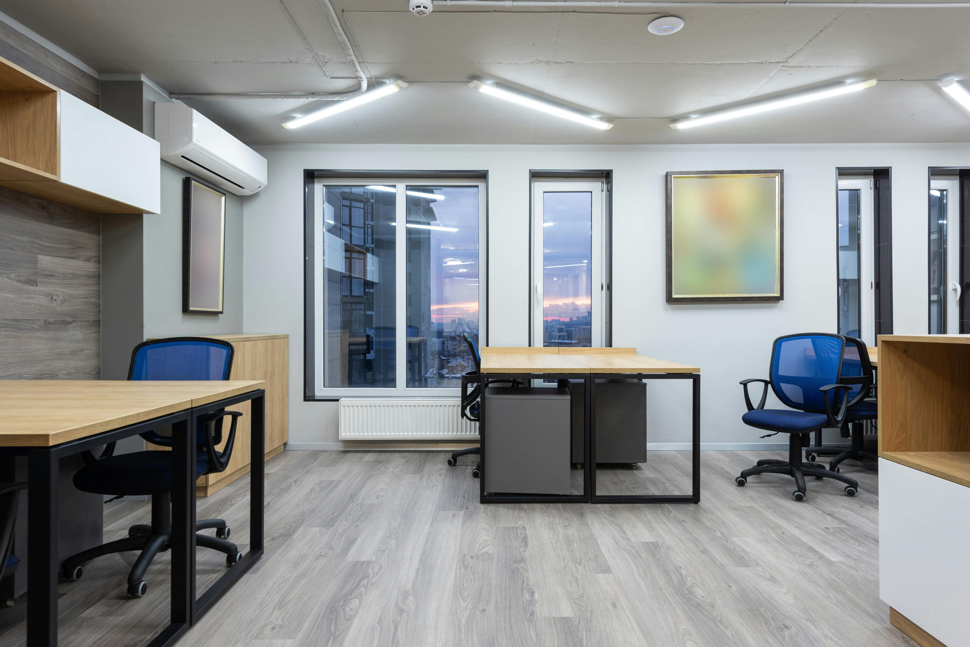 Modern office space with wooden desks, blue chairs, large windows, and abstract art on the wall.