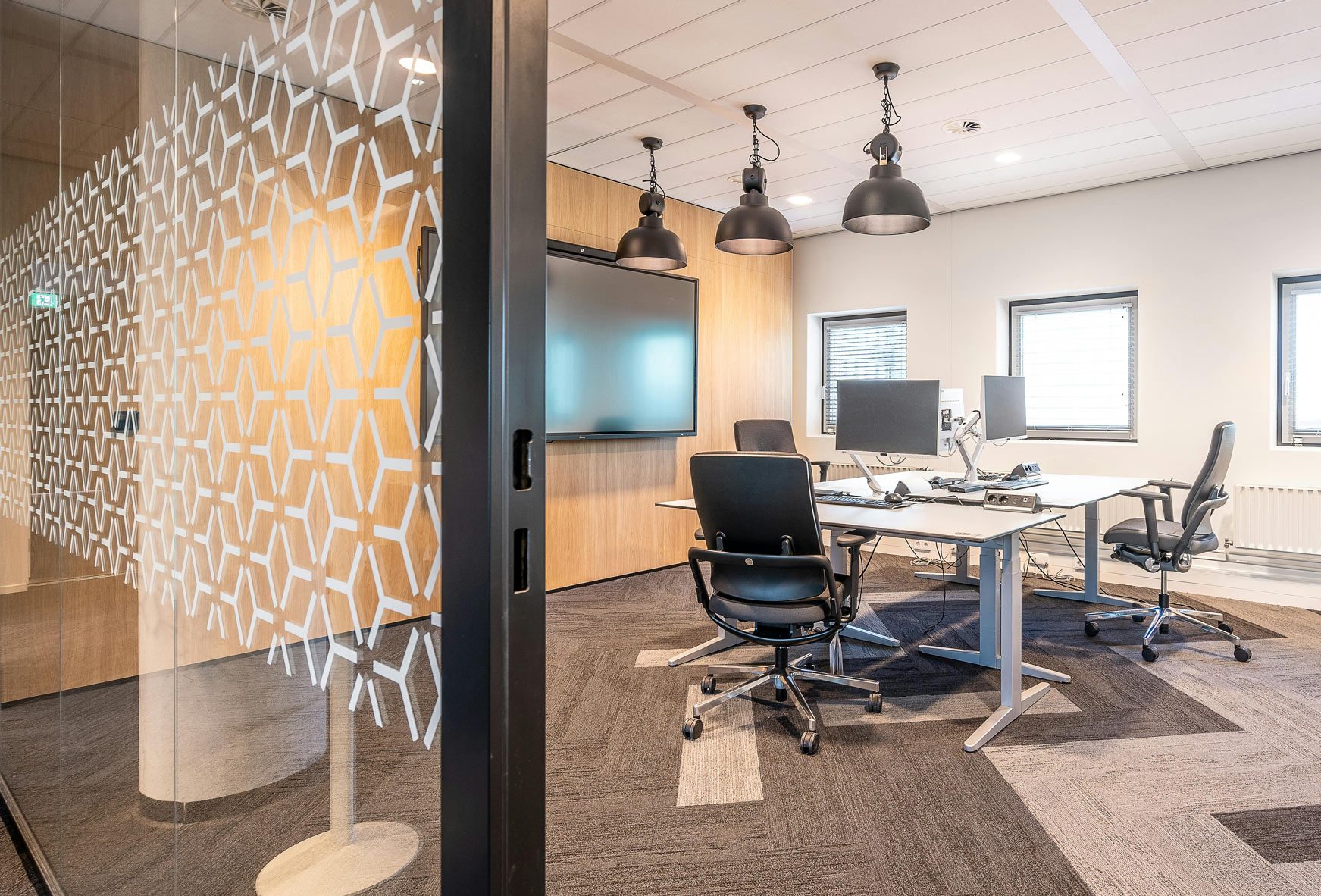 Modern office with two desks, ergonomic chairs, geometric glass door, pendant lights, and a large screen.